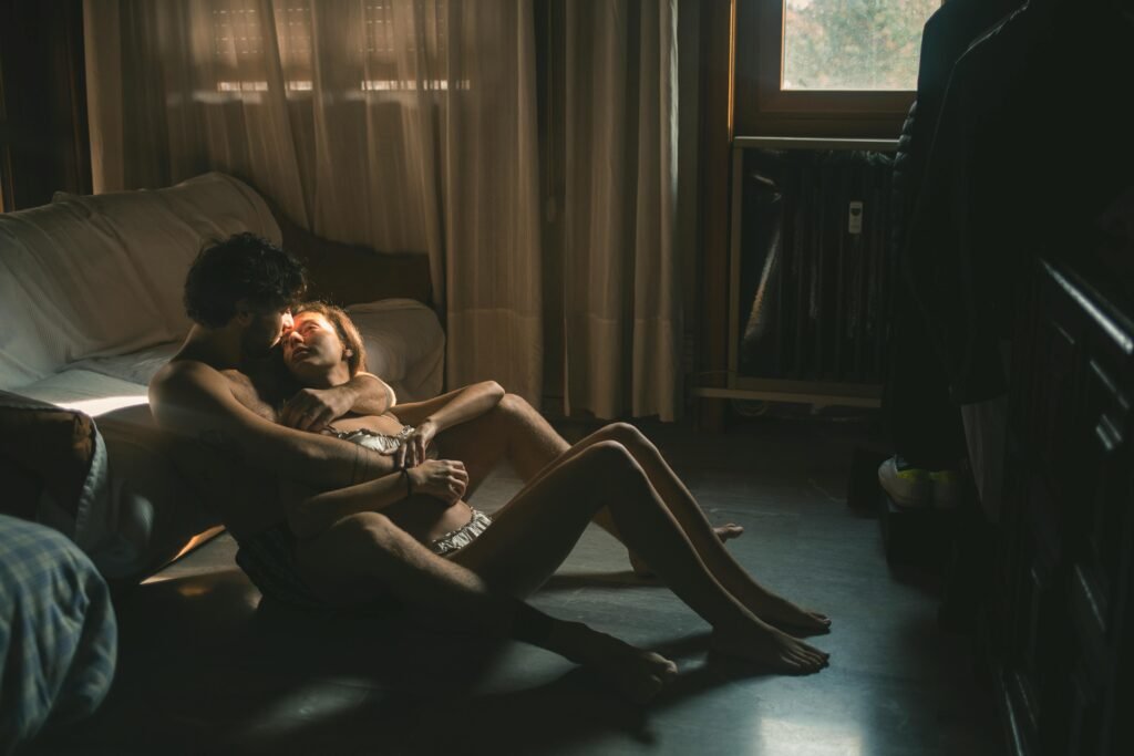 Couple sitting intimately on bedroom floor in warm sunlight streaming through window, showing physical closeness and emotional connection in private intimate moment