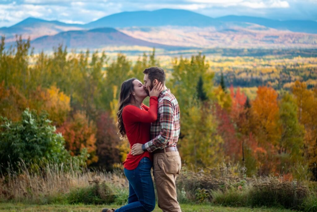 A couple in a monogamous relationship kissing against a breathtaking backdrop of blue mountains and vibrant autumn foliage.