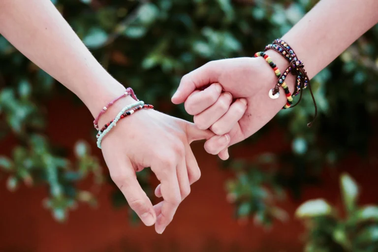 close-up shot of two people's hands linked in a pinky promise against a blurred green and orange background. Both wrists are adorned with several handmade and beaded couple bracelets, emphasizing friendship or romantic connection.