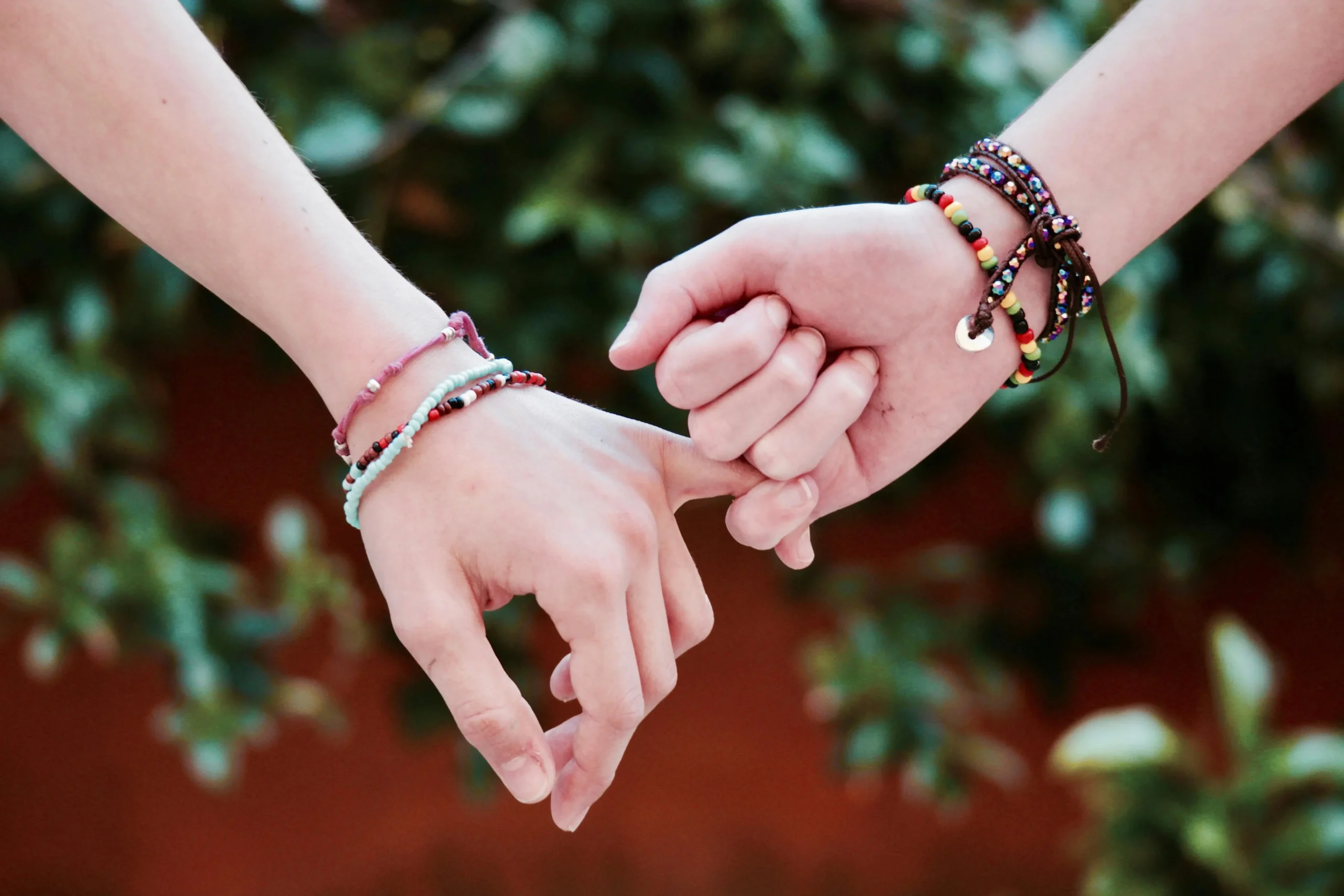 close-up shot of two people's hands linked in a pinky promise against a blurred green and orange background. Both wrists are adorned with several handmade and beaded couple bracelets, emphasizing friendship or romantic connection.