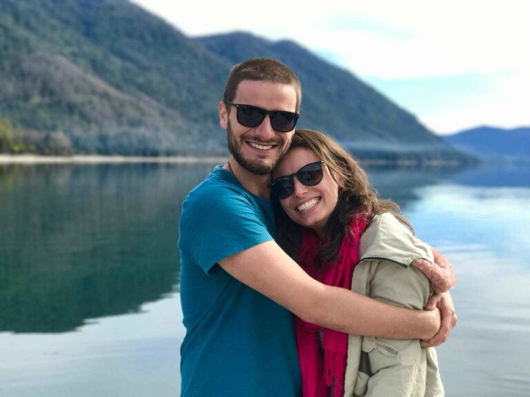 Smiling couple who are having a better relationship after getting Emotional Intimacy Book embracing at scenic mountain lake destination, wearing sunglasses and casual clothes during outdoor adventure