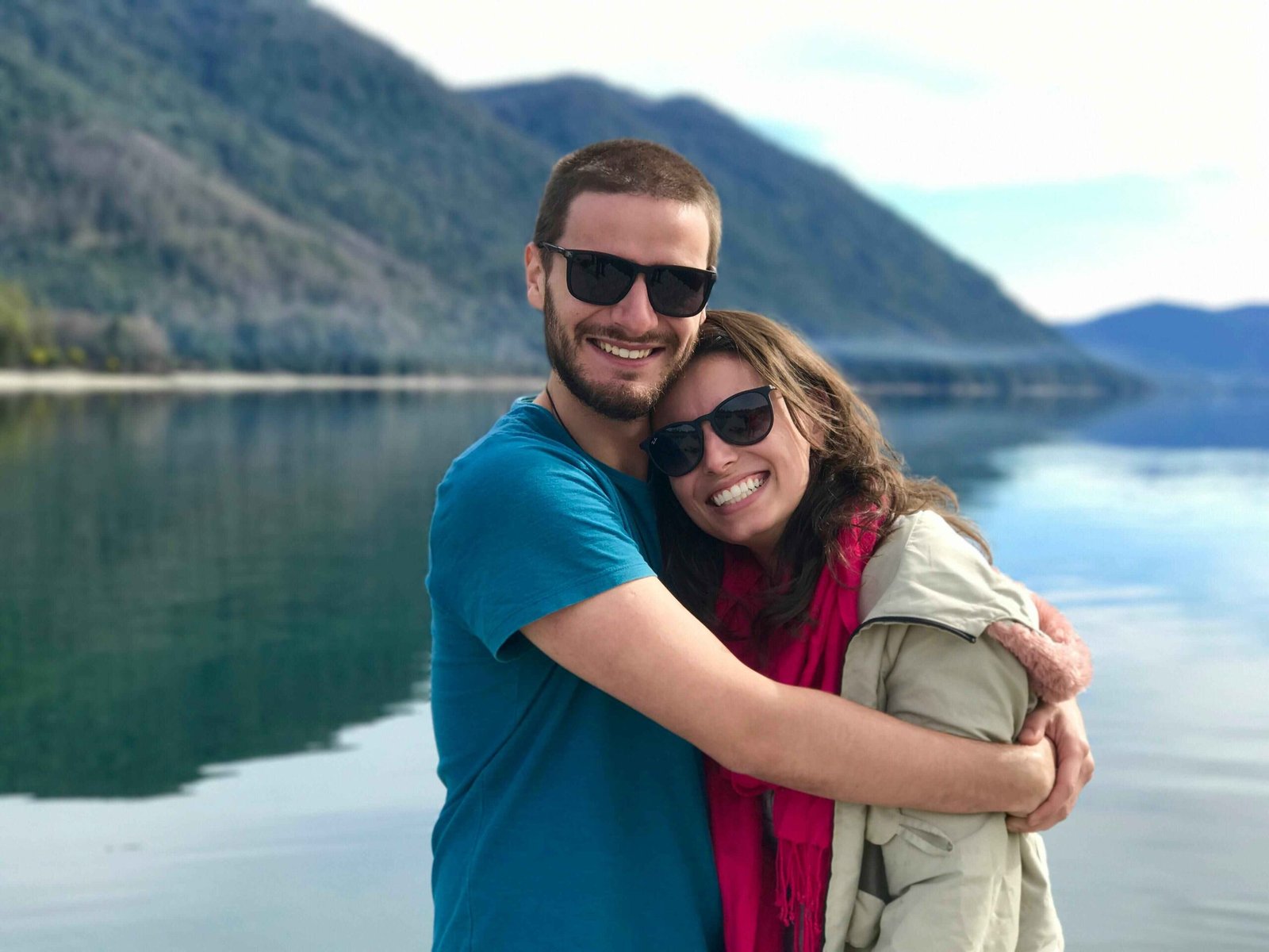 Smiling couple who are having a better relationship after getting Emotional Intimacy Book embracing at scenic mountain lake destination, wearing sunglasses and casual clothes during outdoor adventure