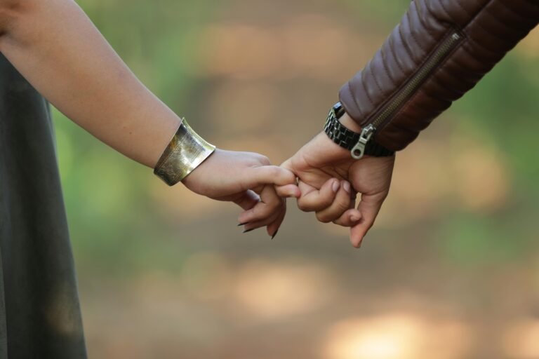 A close-up of two hands making a pinky promise against a blurred green and brown outdoor background. The hand on the left is wearing a wide, hammered gold-tone cuff bracelet. The hand on the right belongs to a person wearing a brown leather jacket with a zippered sleeve and a black wristwatch.