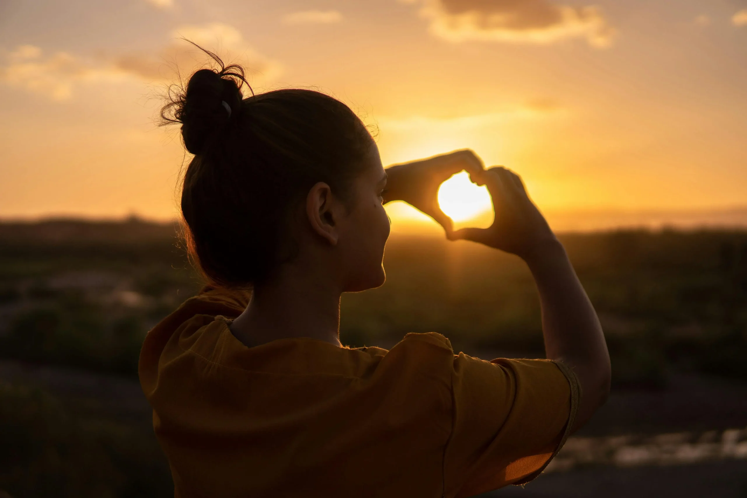 Woman making heart shape with hands at sunset representing how to love yourself and practice self-compassion