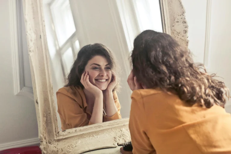 Woman smiling at her reflection in mirror demonstrating how to love yourself through positive self-acceptance