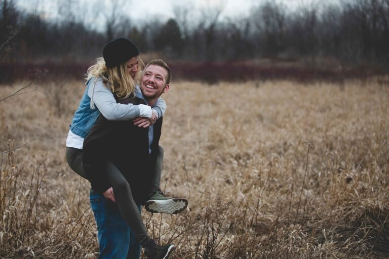 Couple poses in nature field with man giving woman piggyback ride while both laugh authentically during outdoor photo session