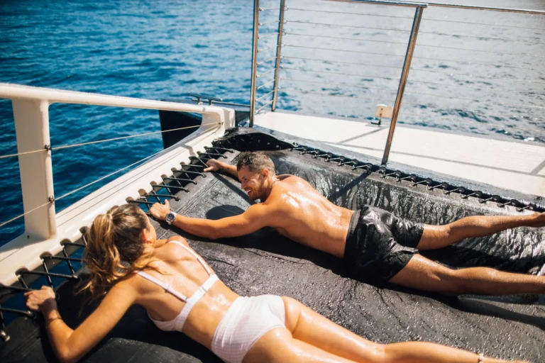 ouple sunbathing on luxury catamaran trampoline deck in Hawaii - man and woman relaxing on boat's mesh net with crystal blue ocean water below