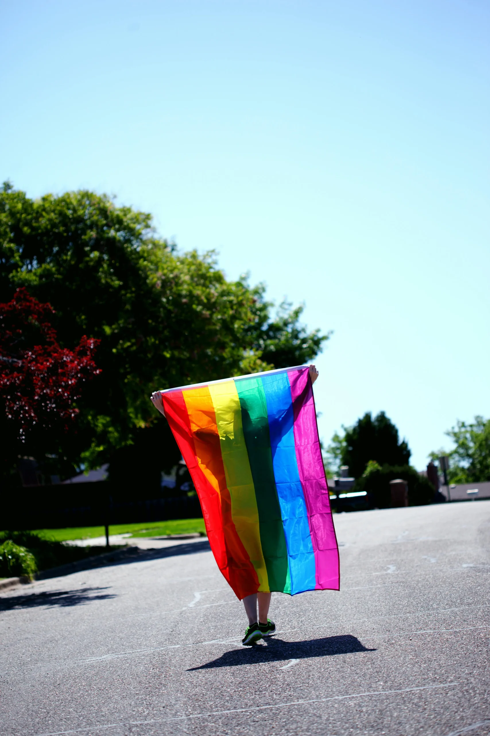 Person draped in vibrant rainbow LGBTQ+ pride flag walking on residential street with trees and blue sky, celebrating love, diversity, and inclusivity in relationships